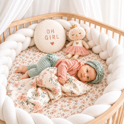 Tour de lit tressé blanc installé dans un lit bébé avec un bébé allongé sur une couverture fleurie.